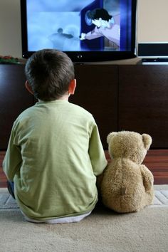 a little boy sitting on the floor watching tv with a teddy bear in front of him