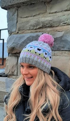 a woman wearing a hat and jacket standing in front of a stone wall with a pink pom - pom on it