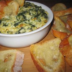 a bowl filled with spinach and bread on top of a wooden table next to slices of bread