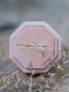 a ring sitting on top of a wooden table next to a flower arrangement and a pink velvet box