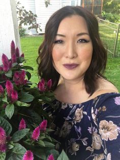 a woman standing next to a bush with purple flowers in front of her and looking at the camera