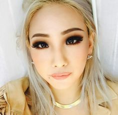 a woman with blonde hair and black eyeliners wearing a tan jacket is looking at the camera