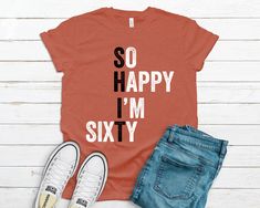 an orange shirt with the words so happy i'm sixty on it next to jeans and sneakers