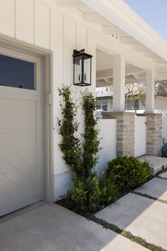 a white house with two doors and some plants on the front porch area, next to it is a light fixture