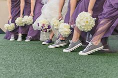 a group of bridesmaids in purple dresses and converse shoes holding bouquets with white flowers