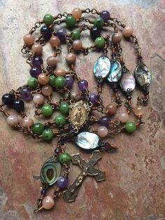 "All wire wrapped handmade rosary with a mix of semi precious gemstones and abalone shell Our Fathers.  I combined 8mm green Canadian Jade, deep violet Amethyst, and creamy rose Sunstone in each decade and Abalone shell ovals accented with amethyst crystals for each Our Father.  There is no other rosary like this.  It is all wire wrapped with 20 gauge coated bronze colored wire (will not fade or rust and no open loops). Double looped split jump rings connect the center and crucifix to prevent th Bohemian Rosary With Natural Stones For Gifts, Spiritual Wire Wrapped Rosary As Gift, Spiritual Gift Wire Wrapped Rosary, Split Jump, Blue Rosary, Handmade Rosary, Amethyst Crystals, Our Father, Miraculous Medal