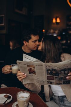 a man and woman kissing while reading a newspaper at a restaurant table with cups of coffee in front of them