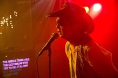 a man with a hat on singing into a microphone in front of a red light