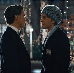 two men standing next to each other in front of christmas lights, one wearing a knitted hat