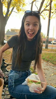 a woman sitting on a park bench holding a sandwich in her right hand and smiling at the camera