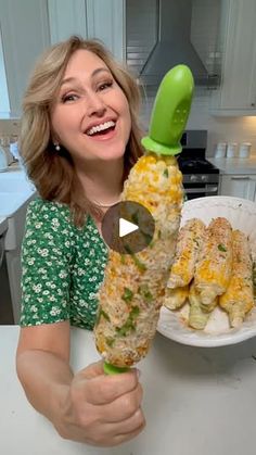 a woman sitting at a table with food in front of her and holding up a corn on the cob