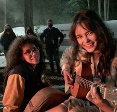 two people sitting on the ground playing guitars in front of snow covered trees and other people