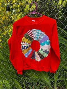 a red sweater with colorful patches hanging on a chain link fence next to yellow flowers