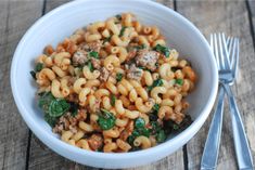 a white bowl filled with pasta and spinach on top of a wooden table next to silverware