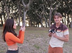 a man and woman standing in the grass taking pictures with their cell phones while one holds a camera