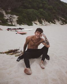 a shirtless man sitting on the beach talking on his cell phone