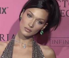 a woman with black hair wearing a silver dress and green earrings on her neck, standing in front of a pink wall