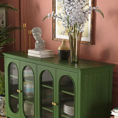 a green cabinet with glass doors and flowers on top in a room decorated with pink walls