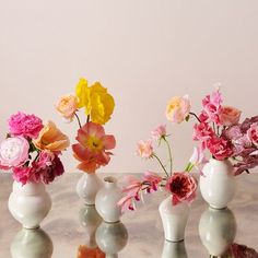 four white vases filled with colorful flowers on top of a marble countertop in front of a pink wall