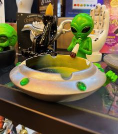 an alien figurine sitting on top of a table next to a bowl filled with liquid