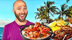 a man holding a plate with food on it in front of the ocean and palm trees