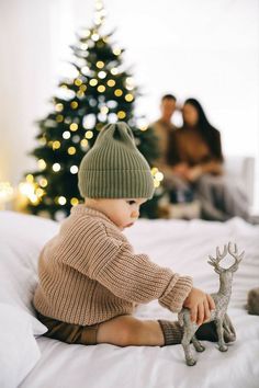 a baby sitting on a bed next to a christmas tree with a toy deer in front of it