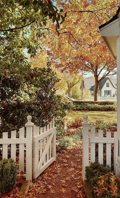 a white picket fence in front of a house with autumn leaves on the ground and trees