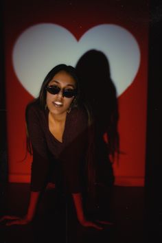 a woman wearing sunglasses is posing in front of a wall with a heart shaped shadow