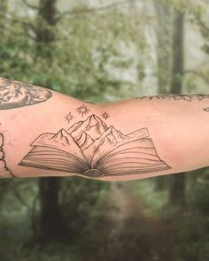 a man's arm with an open book and mountains tattoo on it, in front of some trees