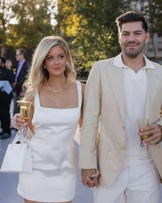 a man and woman walking down the street holding wine glasses in their hands while wearing white outfits