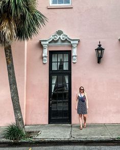 a woman standing in front of a pink building with black doors and palm trees on the sidewalk