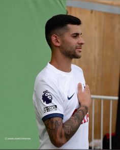 a man with tattoos standing in front of a green wall and wearing a white shirt