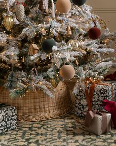 a christmas tree with presents under it in front of a basket on the floor next to two wrapped gifts