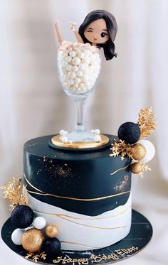 a black and gold birthday cake with a doll on top that is holding a glass filled with candy