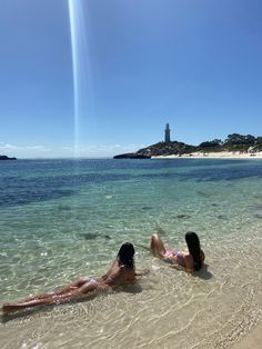 two women laying on their stomachs in shallow water near an island with a light house in the distance