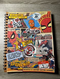 a spiral notebook covered in comic stickers on top of a wooden table next to a cup of coffee