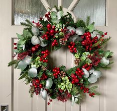 a christmas wreath with red berries, greenery and pine cones hanging on a door