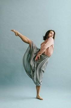 a woman standing on one leg with her legs spread out in the air, wearing a pink shirt and gray skirt