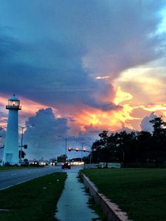 the sun is setting behind a light house on the side of the road with grass in front of it