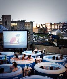 people are sitting in large tubs watching movies on the big screen outside at dusk