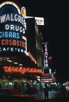 people are walking on the sidewalk in front of some neon signs and buildings at night