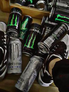 several cans and cans of monster energy drink sitting on top of a wooden table with one person's feet in the air