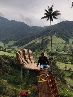 a man sitting on top of a giant hand sculpture in the middle of a lush green valley