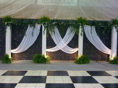 an outdoor wedding setup with white drapes and greenery on the wall, surrounded by black and white checkered flooring