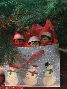 three elfs are sitting in a bag under the christmas tree, with snowmen on it