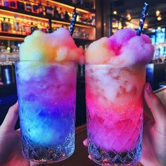 two people holding up colorful drinks in front of a bar