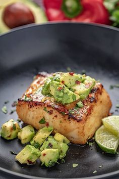 a piece of fish with avocado and lime slices on the side in a black plate