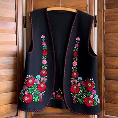 a black vest with red and pink flowers is hanging on a wooden hanger in front of a door