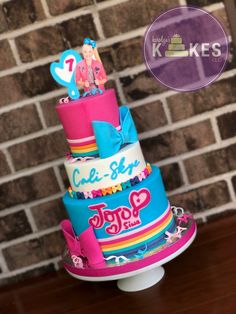 a three tiered cake with pink, blue and white frosting on top sitting on a table in front of a brick wall