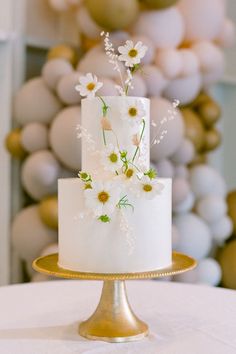 a white wedding cake with daisies and greenery on top sits on a gold plate